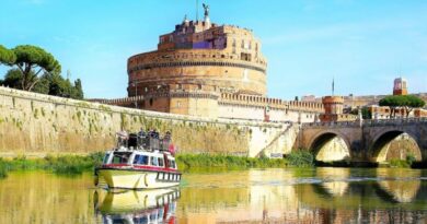 Battello sul Tevere davanti a Castel Sant’Angelo a Roma in una giornata di sole, con riflessi sull’acqua e ponte sullo sfondo.