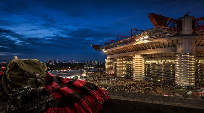 San Siro illuminato di sera durante una partita del Milan, con tifosi rossoneri sugli spalti, sciarpa rossonera, zaino verde e macchina fotografica in primo piano.